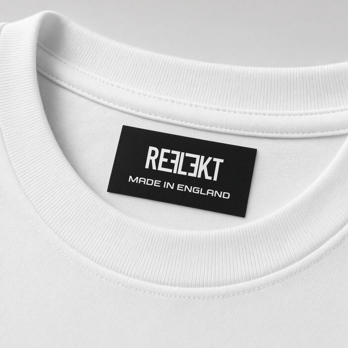 Close-up of a white crewneck t-shirt showing the inner neck label with the brand name 'REFLEKT MADE IN ENGLAND' printed in white on a black background.
