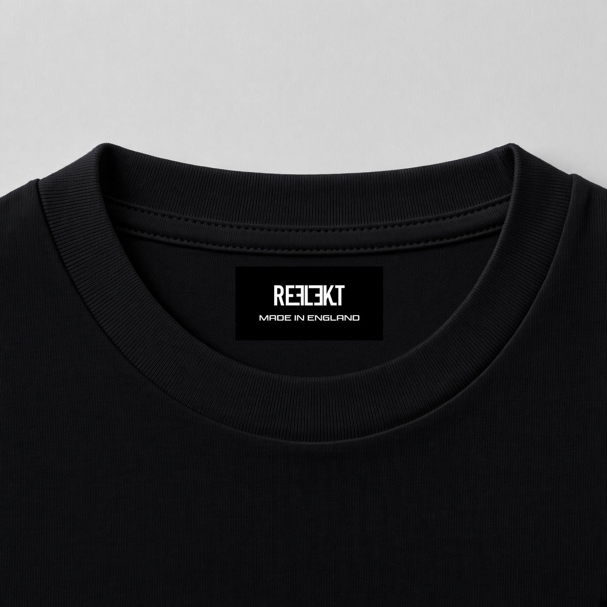 Close-up of a black crewneck t-shirt showing the inner neck label with the brand name 'REFLEKT MADE IN ENGLAND' in white text, set against a light gray background.