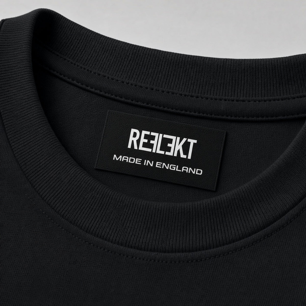 Close-up of a black crewneck t-shirt showing the inner neck label with the brand name 'REFLEKT MADE IN ENGLAND' in white text, set against a light gray background.