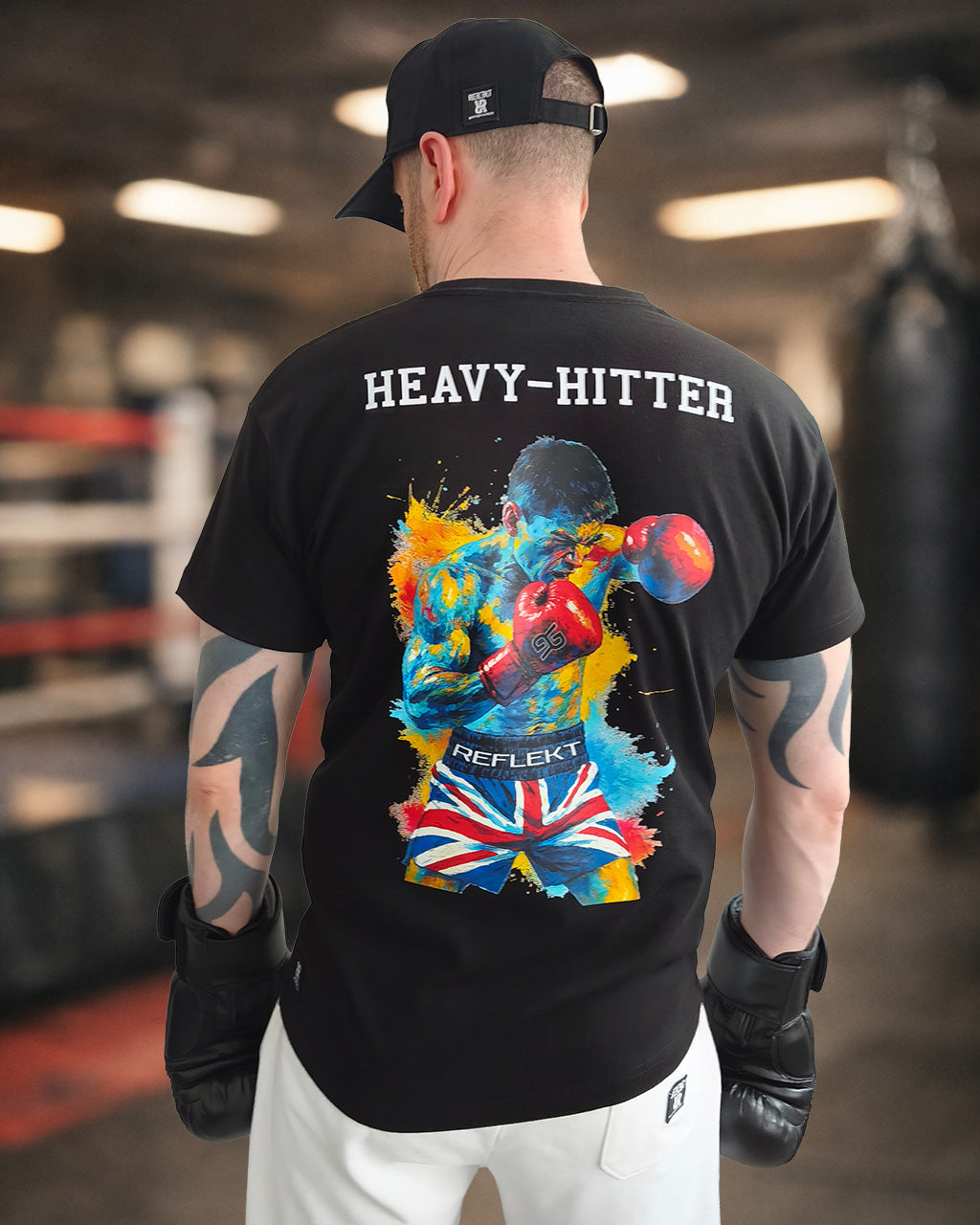 A man stands in a boxing gym, facing away from the camera. He wears a black cap, black gloves, a black T‑shirt, and white trousers. The T‑shirt features a vivid graphic of a boxer mid‑punch, surrounded by splashes of blue, yellow, and red paint. The boxer in the design wears red gloves and Union Jack shorts with the word “REFLEKT” on the waistband. Above the artwork, the shirt reads “HEAVY‑HITTER.” The blurred background shows a boxing ring and punching bag, creating a dynamic, athletic atmosphere.