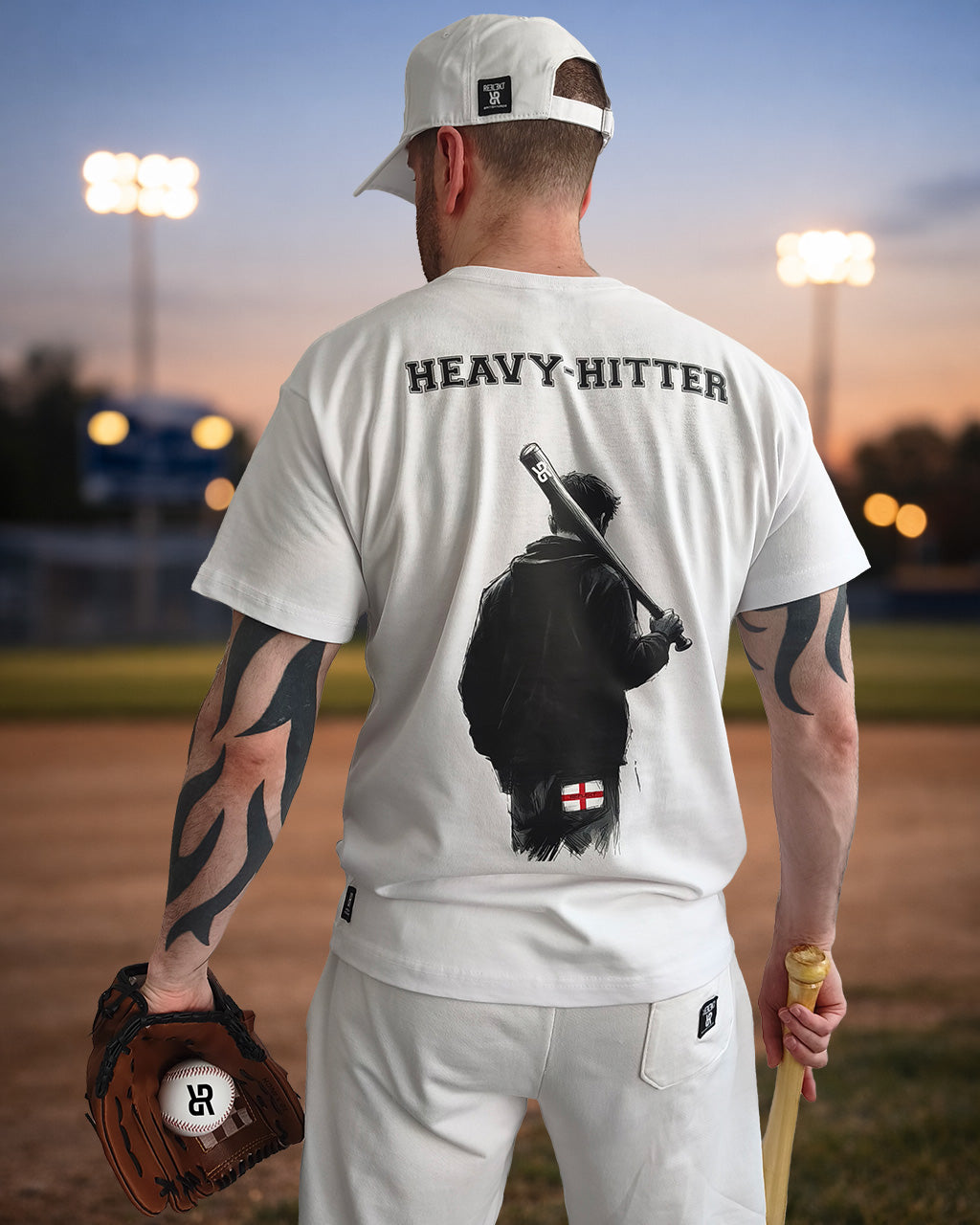 A man stands on a baseball field at dusk, facing away from the camera. He wears a white cap, white T‑shirt, and white trousers. The back of his shirt reads “HEAVY‑HITTER” above a graphic of a figure holding a bat with a small England flag near the bottom. He holds a baseball glove in one hand and a bat in the other, with stadium lights glowing softly in the background.