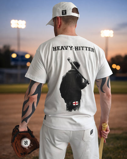 A man stands on a baseball field at dusk, facing away from the camera. He wears a white cap, white T‑shirt, and white trousers. The back of his shirt reads “HEAVY‑HITTER” above a graphic of a figure holding a bat with a small England flag near the bottom. He holds a baseball glove in one hand and a bat in the other, with stadium lights glowing softly in the background.