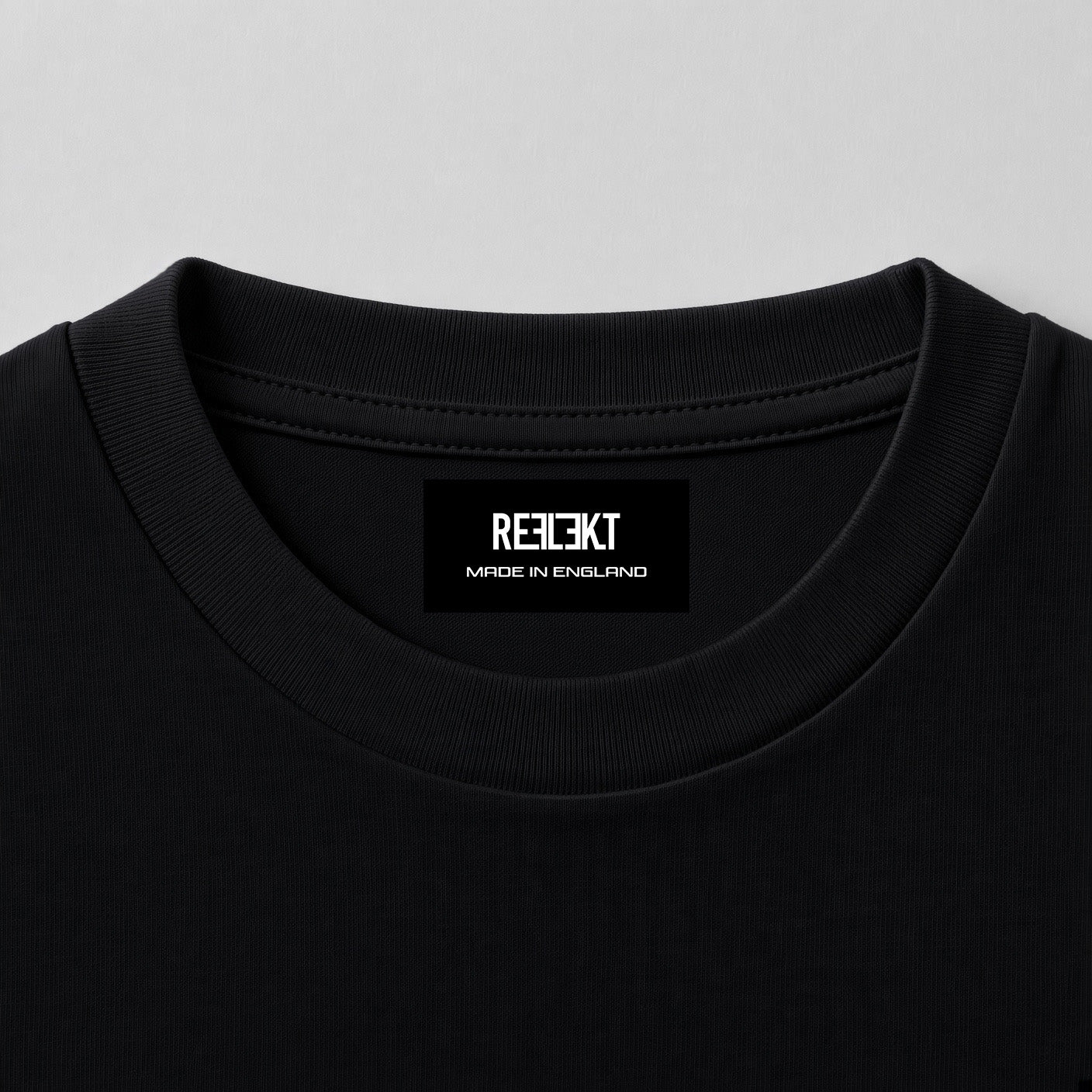 Close-up of a black crewneck t-shirt showing the inner neck label with the brand name 'REFLEKT MADE IN ENGLAND' in white text, set against a light gray background.