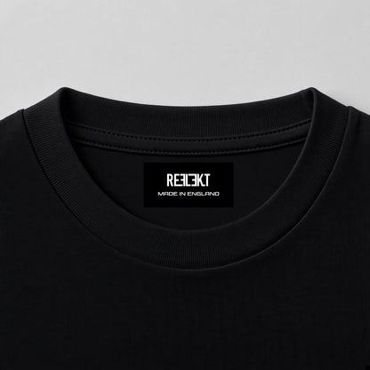 Close-up of a black crewneck t-shirt showing the inner neck label with the brand name 'REFLEKT MADE IN ENGLAND' in white text, set against a light gray background.