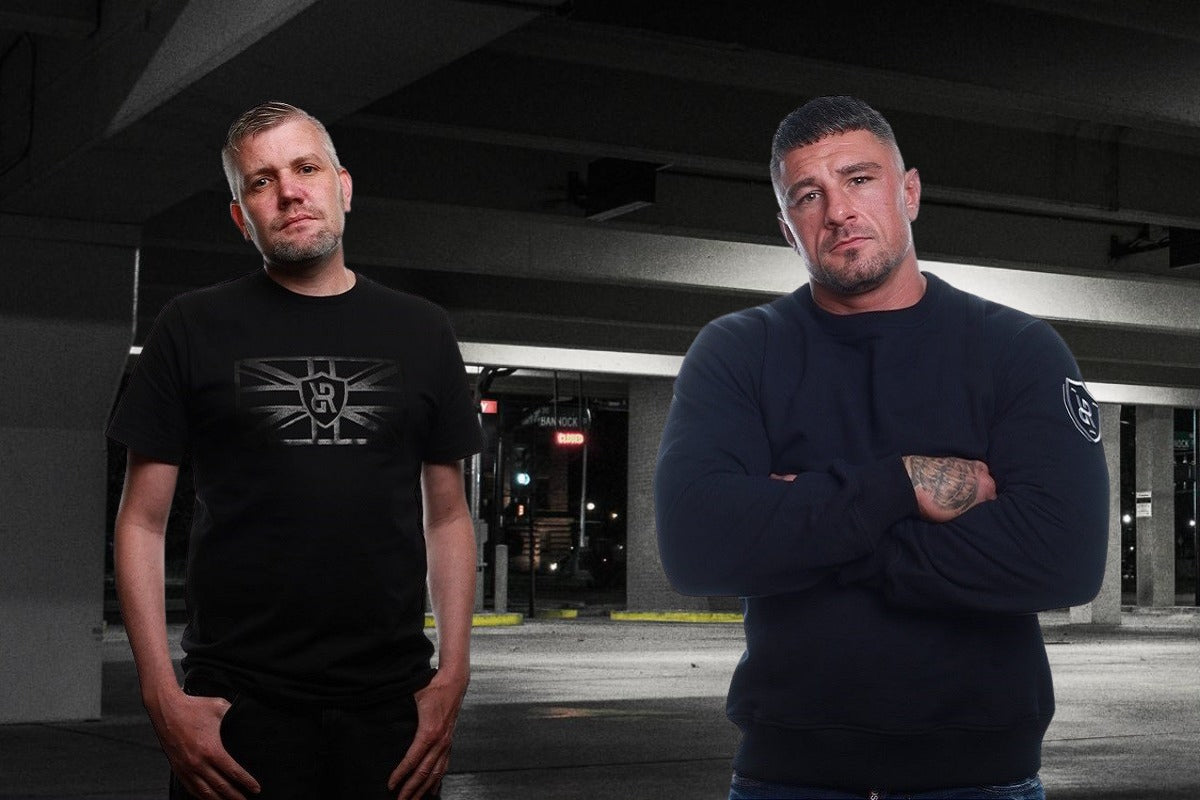 Two men standing in a parking garage, one wearing a black t-shirt with a logo and the other in a dark sweater.